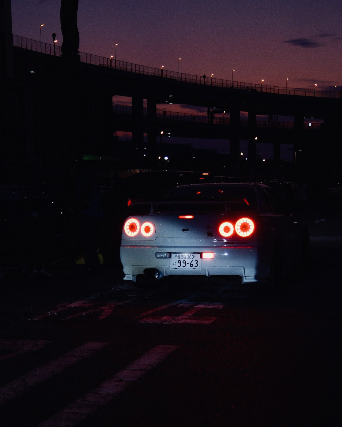 Nissan Skyline R34 GT-R rear taillights glowing under a Tokyo expressway at dusk