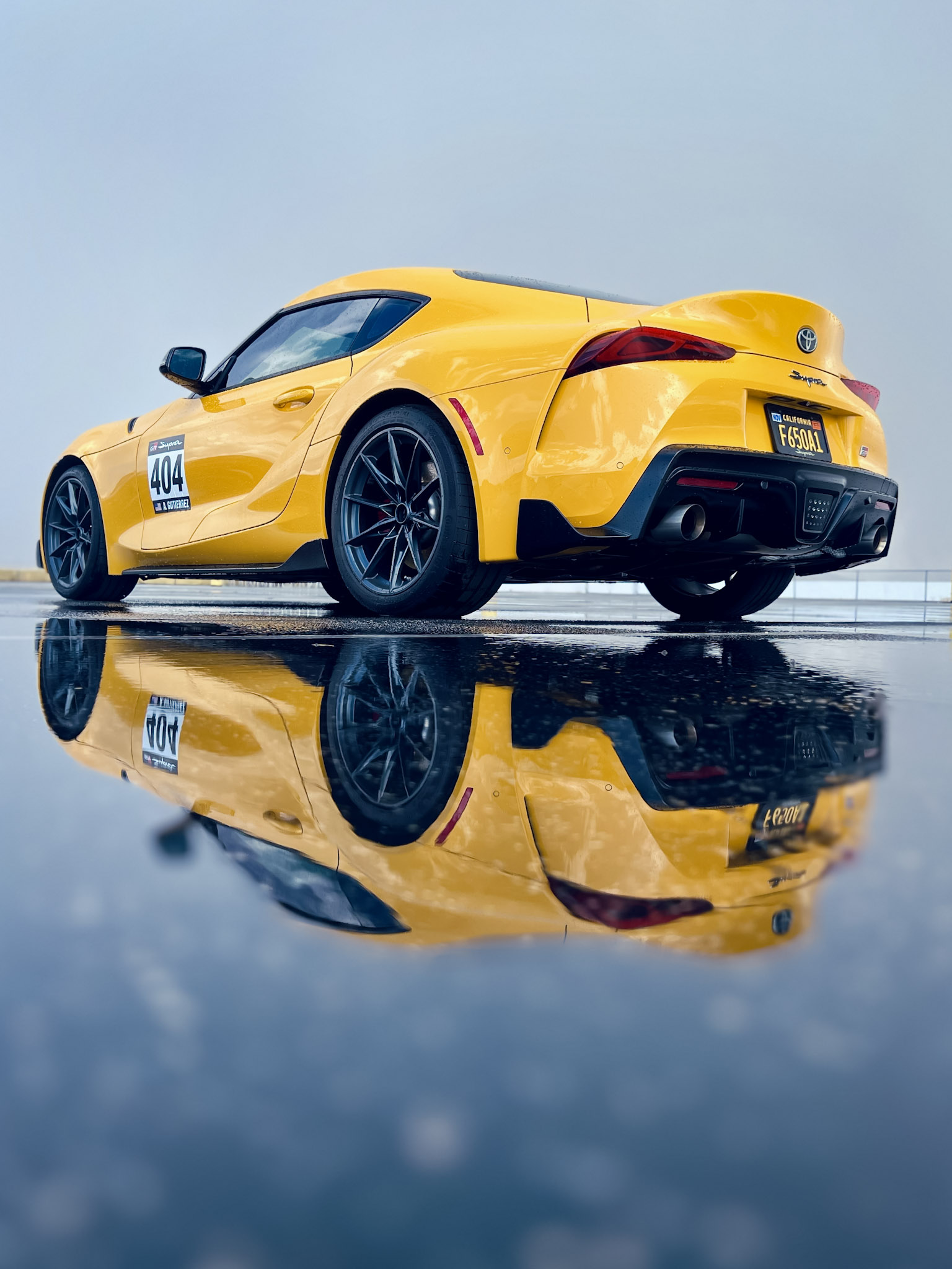 Yellow Toyota GR Supra #404 perfectly reflected in wet pavement on a grey morning